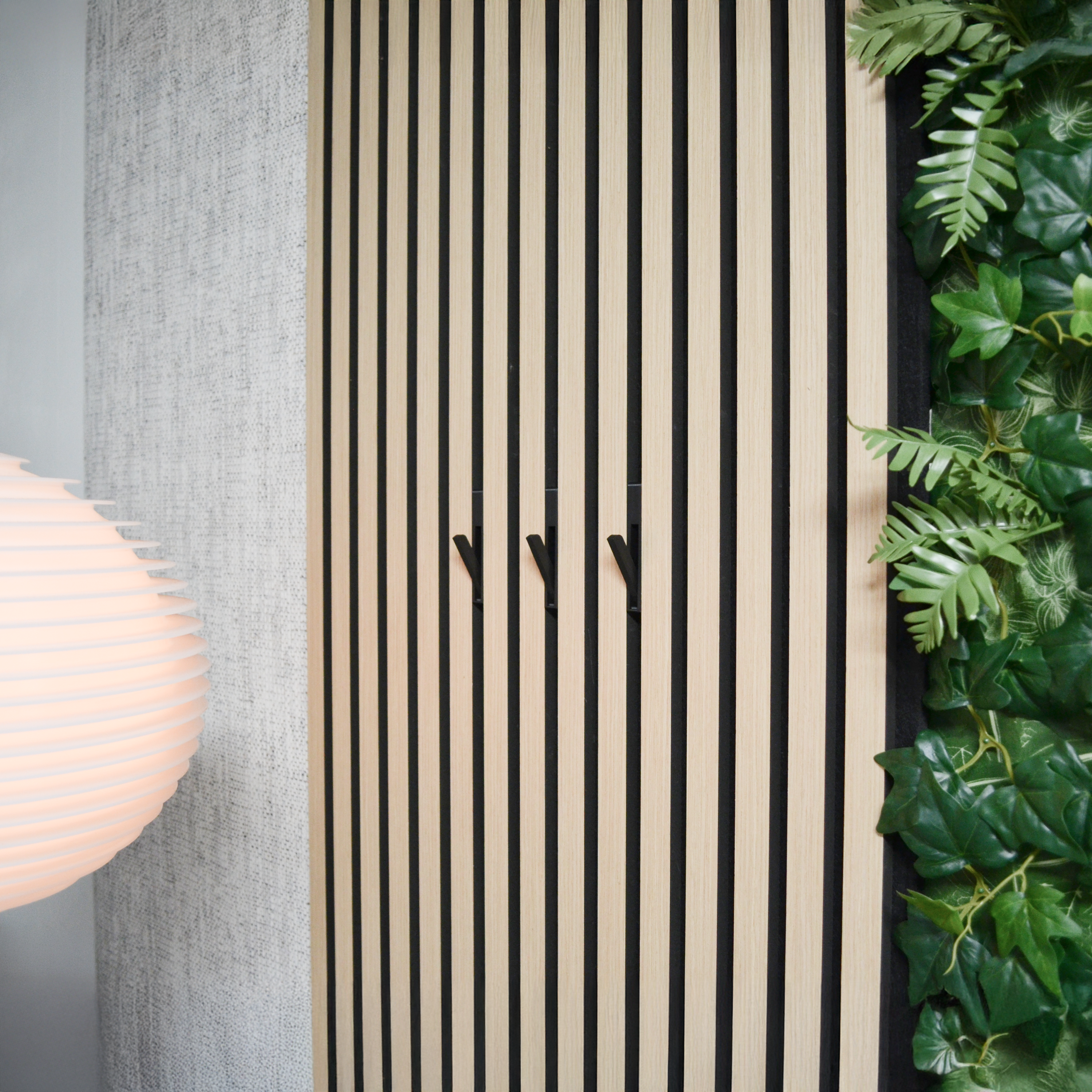 Modernes Wandpaneel mit vertikalen Holz- und schwarzen Akustikstreifen, ausgestattet mit klapphaken "HIDE". Drei geöffnete Haken sind sichtbar. Dekoriert mit grünen Pflanzen und einer Designerlampe. Hochwertiges Akustikpaneel zubehör für stilvolle Garderoben mit bohrfreier Montage.