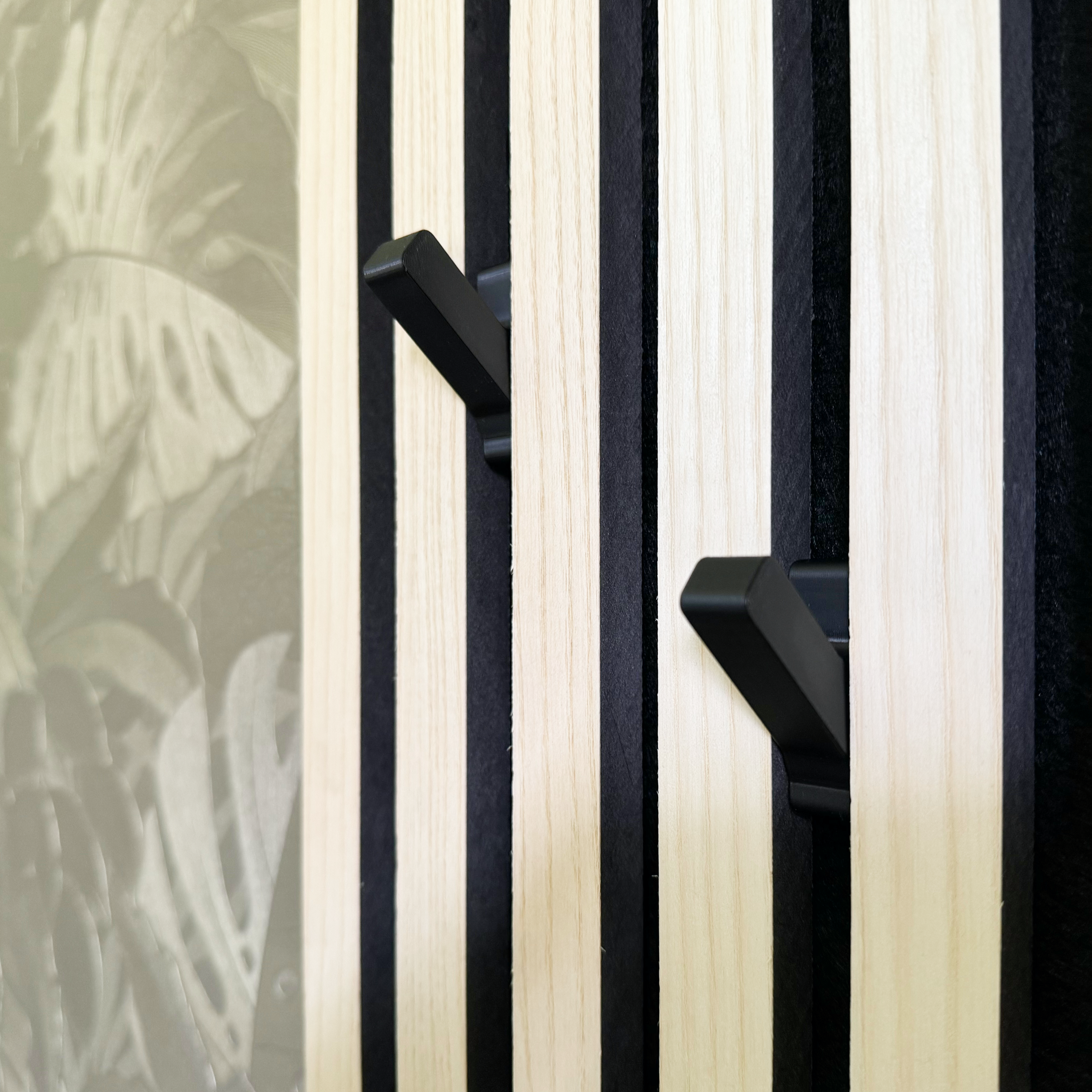 Schwarze Design-Haken als praktisches Akustikpannel Zubehör, montiert auf modernen Wandpaneelen in Holzoptik mit schwarzen Fugen. Ideal für die stilvolle Gestaltung einer Garderobe ohne Bohren. Die Haken passen perfekt in Akustikpaneele und Wandpaneele und bieten eine flexible, ästhetische Lösung zur Aufbewahrung von Kleidung und Accessoires. Funktionalität trifft auf minimalistisches Design.