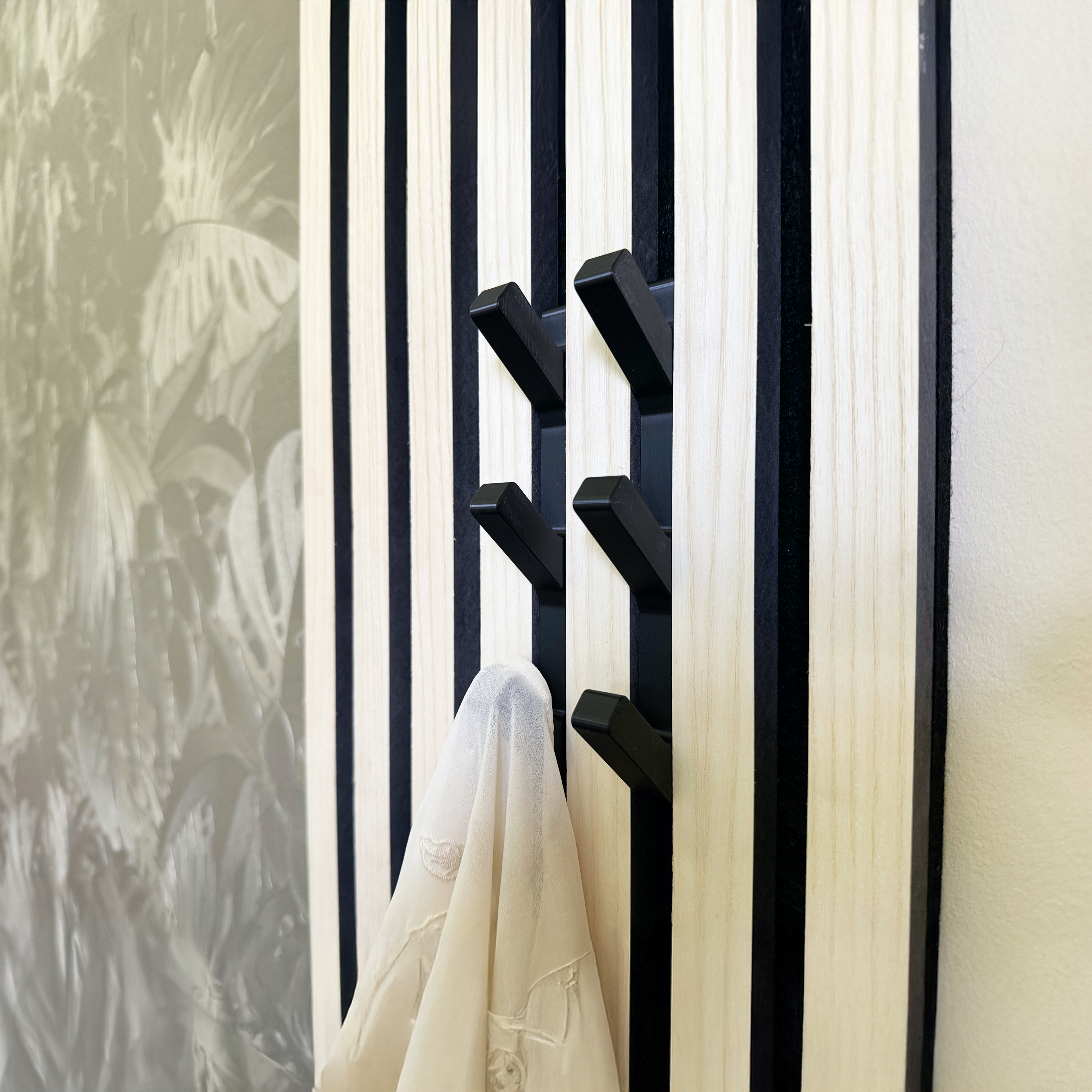 Moderne Wandpaneele kombiniert mit Akustikpaneel Zubehör, ideal als Garderobe. Vertikale Holzlatten in hellem Ton mit schwarzen Streifen zur Schallabsorption. Schwarze Haken bieten funktionalen Stauraum, z. B. für Kleidung. Perfekte Kombination aus ästhetischem Design und praktischer Nutzung für Wohn- oder Büroräume.