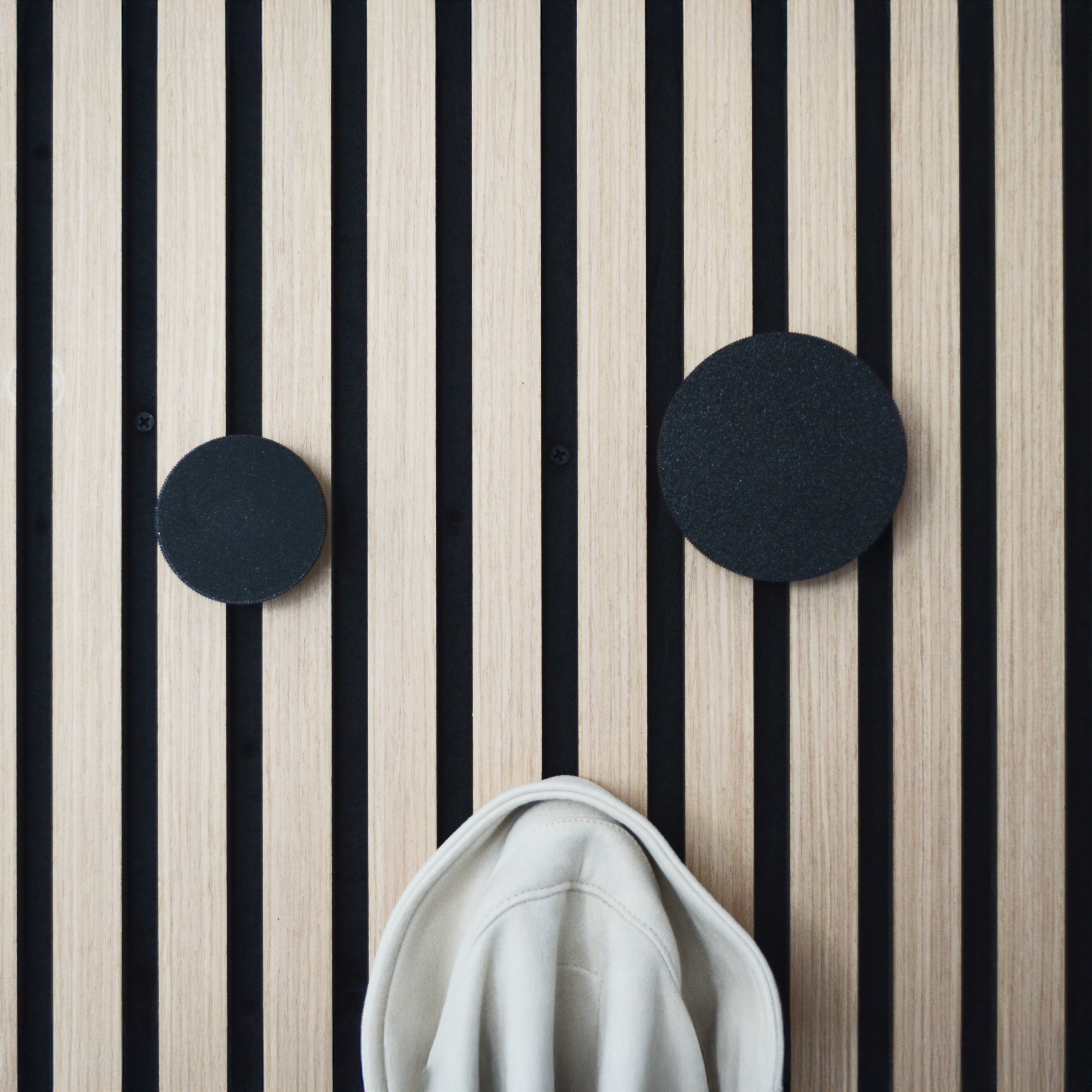 Modernes Wandpaneel mit vertikalen Holz- und schwarzen Akustikstreifen, ausgestattet mit runden, schwarzen Wandhaken. Ein beigefarbener Hoodie hängt an einem Haken. Hochwertiges Akustikpaneel zubehör für stilvolle Garderoben, ideal für eine bohrfreie Montage in Flur oder Eingangsbereich.