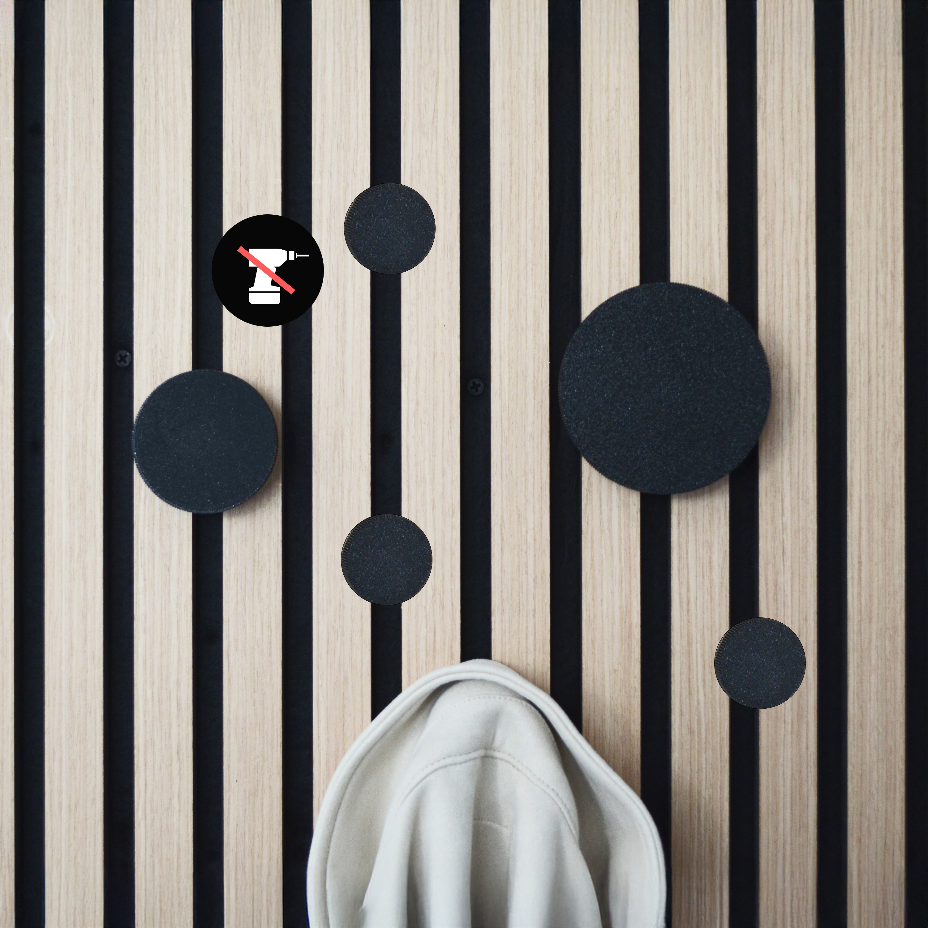 Modernes Wandpaneel aus Holz mit schwarzen Streifen und runden, schwarzen Haken in verschiedenen Größen. Ein beigefarbener Hoodie hängt daran. Ein Symbol mit durchgestrichenem Bohrer deutet auf eine bohrfreie Montage hin. Stilvolle Wandgarderobe für Flur & Eingangsbereich.
